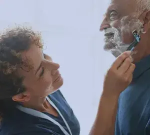 Caregiver helping an elderly man shave with a razor, both smiling.